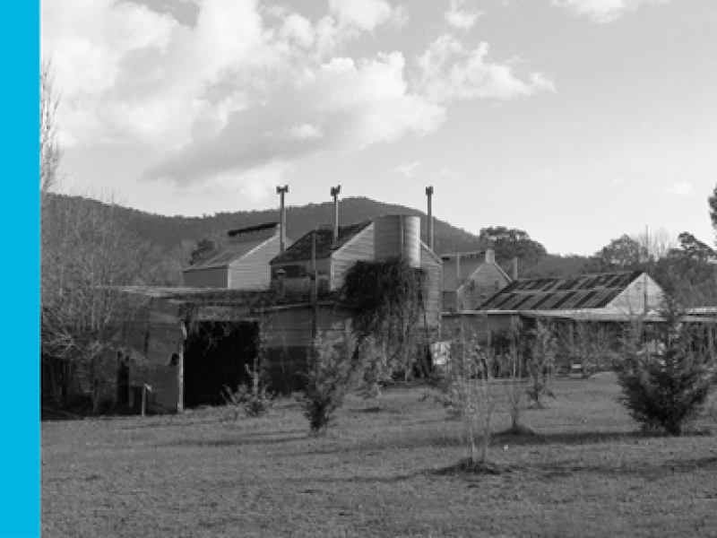 A black and white image of a corrugated iron building with hills in the background.