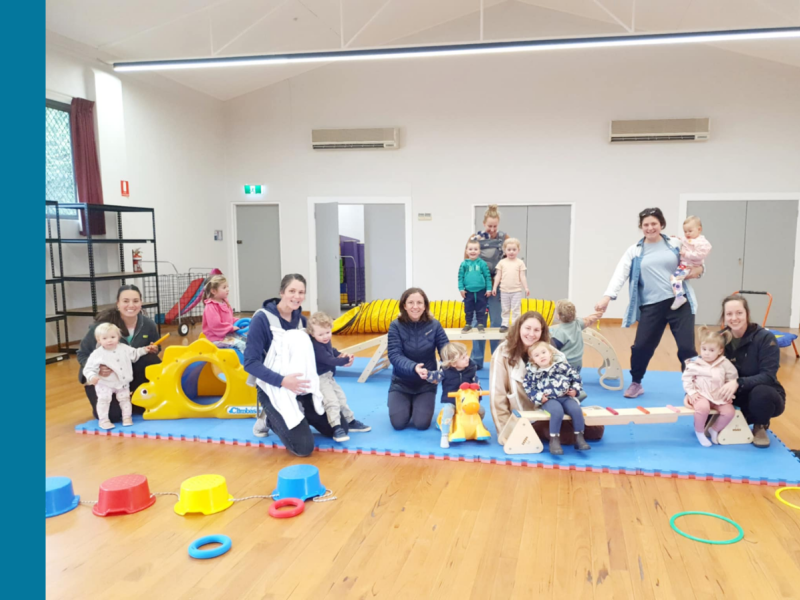 Seven mothers are in a hall with their children with toys scattered around them.