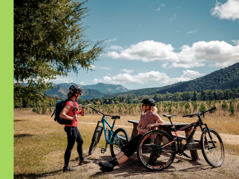 Two women are together with pushbikes. One is standing, while the other woman is sitting at a picnic table. There are hills in the background.