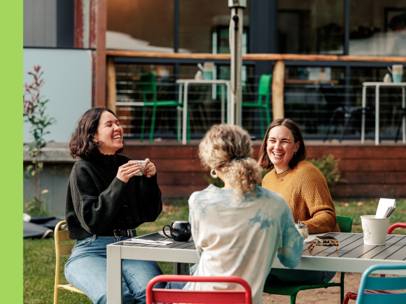 Three women sit at a cafe table outside. The woman on the left is wearing blue jeans and a green jumper, laughing while she is holding a cup of tea. The woman to the back is wearing an orange jumper and is smiling at the woman facing away, who is wearing a tie-dyed shirt.