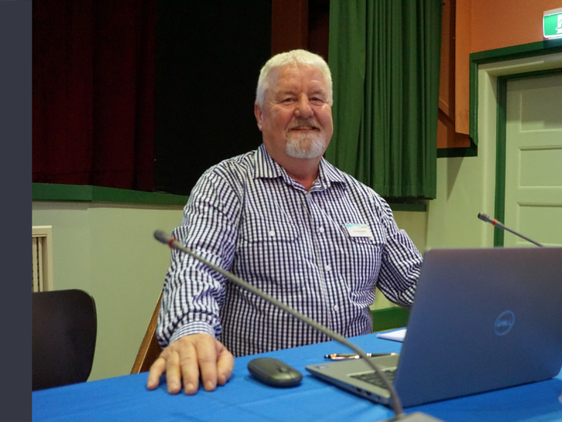 A middle-aged white man with white hair sits behind a desk, which has a laptop and a mouse on it. He is smiling at the camera.