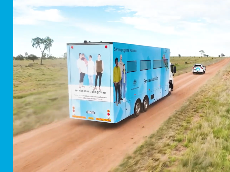 A light blue truck drives along a red dirt road with flat green land on either side. The truck is a mobile service centre.