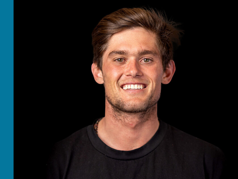 A young white man with brown hair and a black shirt is smiling at the camera.
