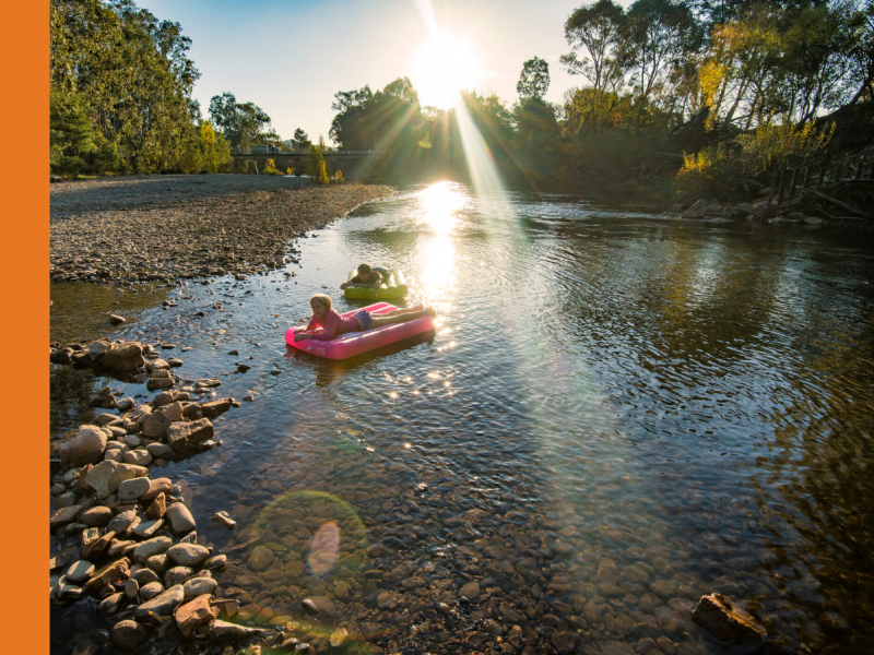 Two young people floating on lilos in the river with the Nimmo Bridge in the background.