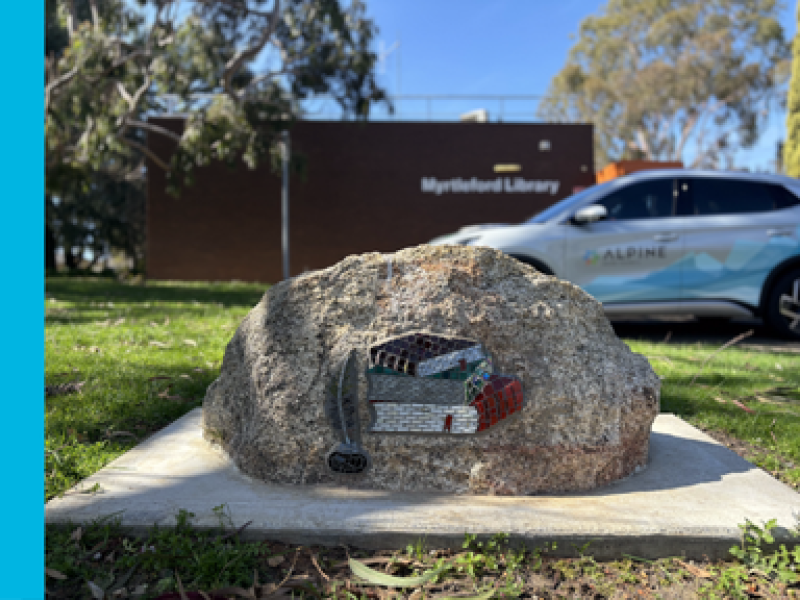 A rock with mosaic books on it is in front of a brick building that says Myrtleford Library on it.