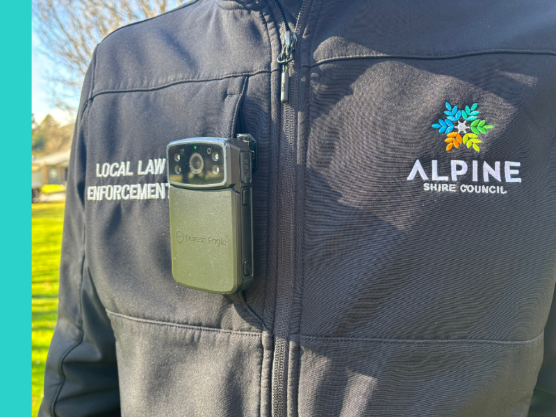 A person wearing a black jacket that says Alpine Shire Council on the right side of the chest and Local Laws Enforcement on the left side. They have a camera clipped to the chest pocket.
