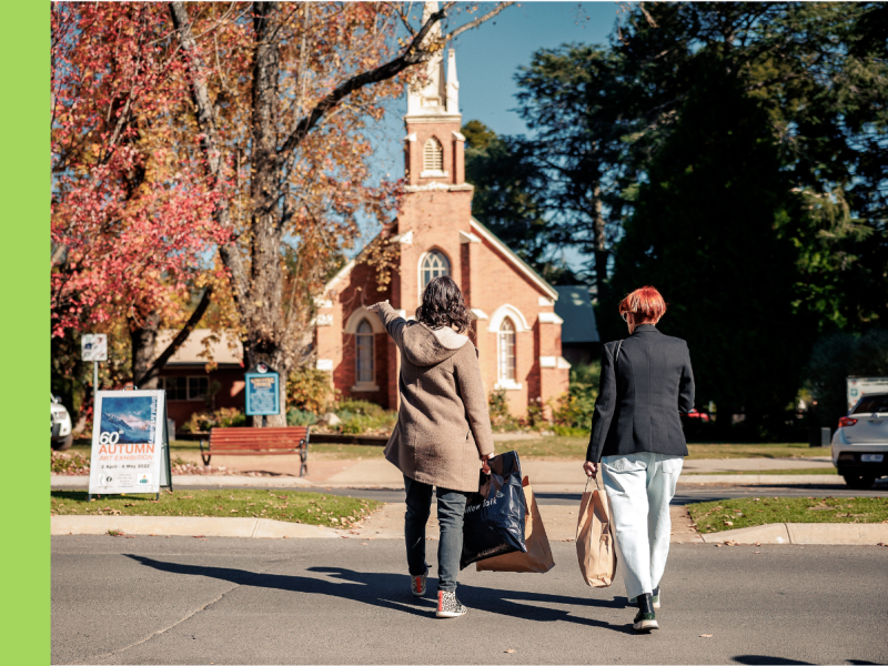 Two people walk across a road in front of a red brick church with a tall spire at the front. There are autumn trees next to the church with red leaves.