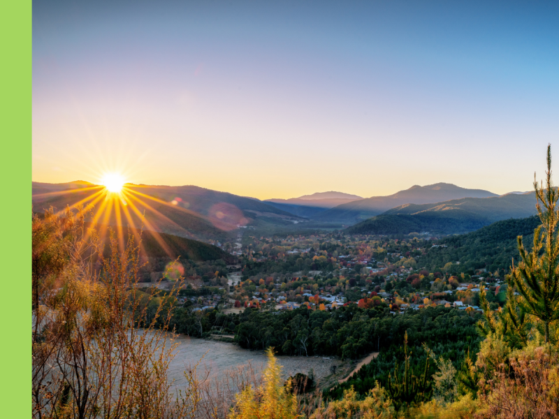 A valley with a sun flare over the hills and autumn trees dotted amongst the houses. There is a big body of water in front of the town.
