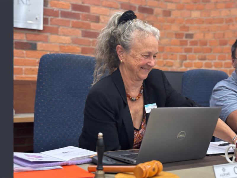 A woman sits at a desk in front of an open laptop. She is reaching over the desk and smiling.