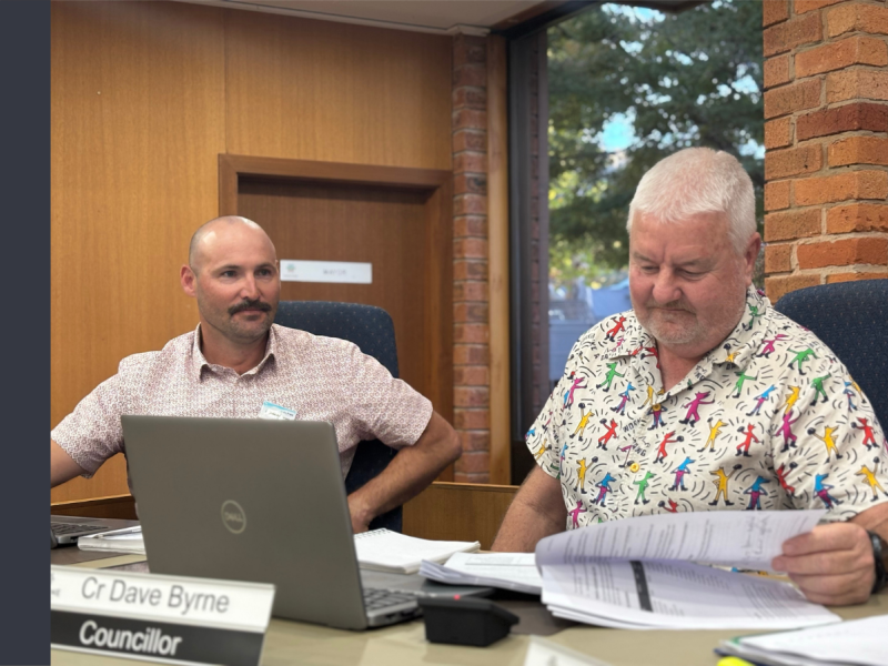 Two white men sit behind a desk. There is a nameplate in front of the man on the right that reads Cr Dave Byrne, Councillor.