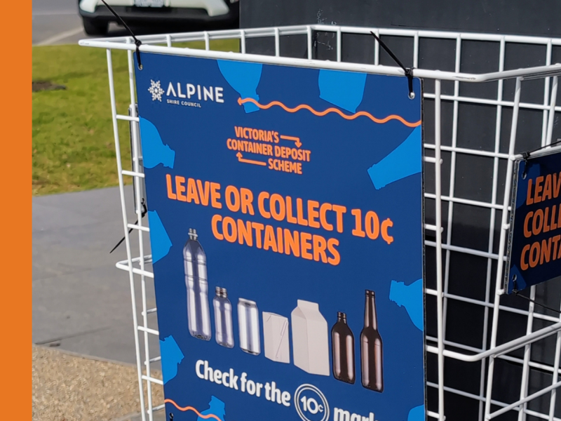 A white, wire basket attached to the side of a public recycling bin, with a blue and orange sign saying 'Leave or collect 10c containers'