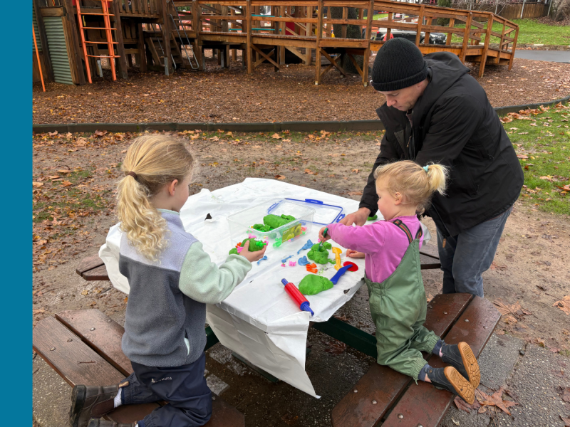 A father stands next to an outdoor picnic table with a playground structure in the background. His two young daughters are kneeling on the table benches painting on a large sheed of paper with their dad's help.