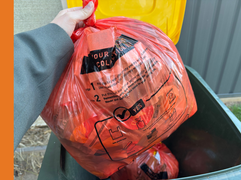 A person places an orange bad in a yellow-lidded recycling bin.