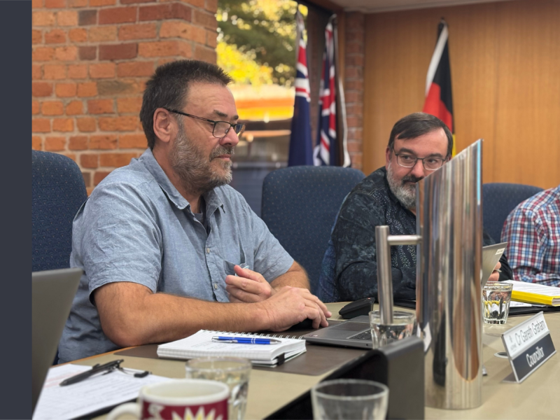 A white man with dark hair and glasses sits behind a desk, looking at a laptop. Another man with dark hair and a white beard sits next to him, looking in his direction.