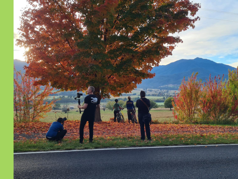 Three people with cameras stand on the side of a road, with two people standing next to bikes in front of a mountain.