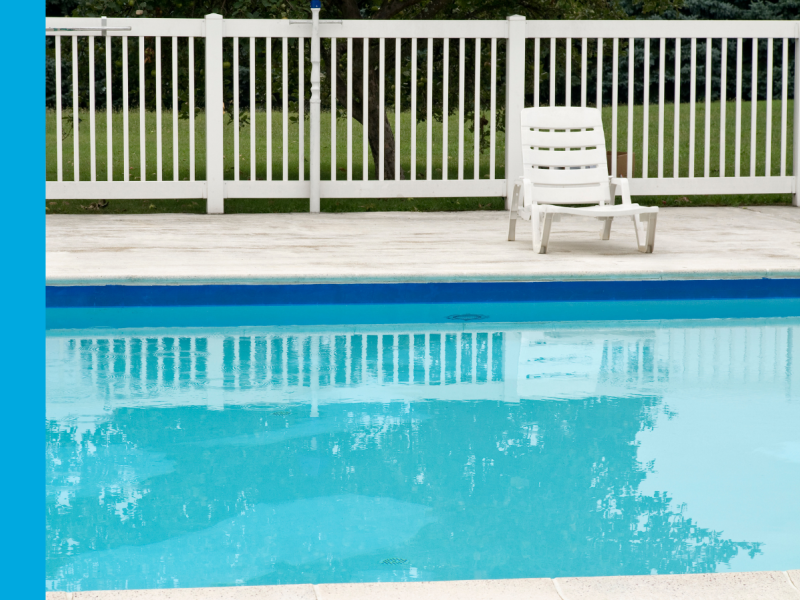 A swimming pool surrounded by a white fence with a lounge chair nearby.