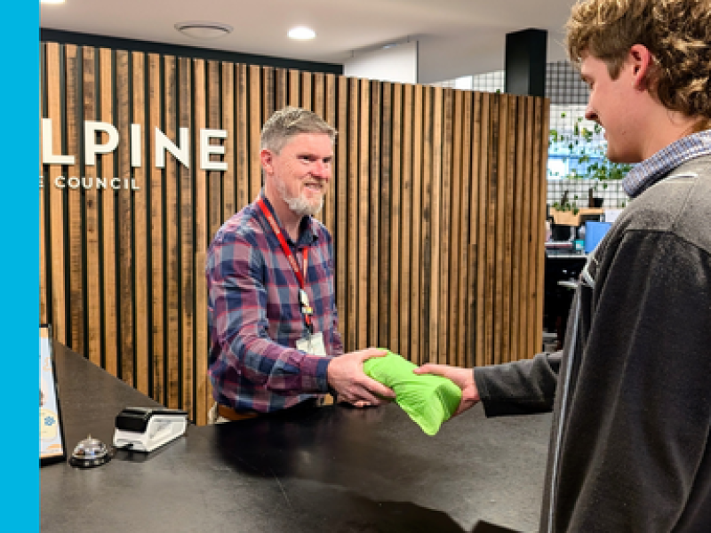 A male customer service staff member hands a roll of green FOGO bags across the counter to a young male with blond hair wearing a dark grey jacket.