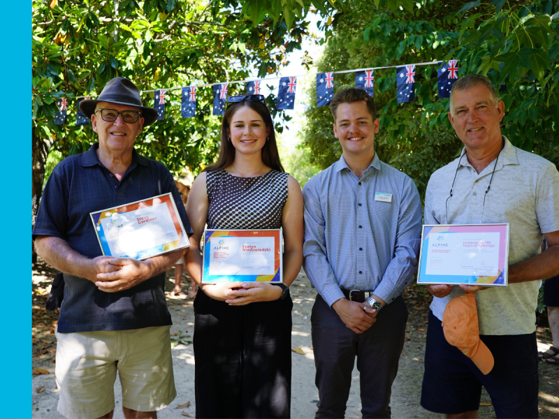 Three men and a woman stand in a row in a park with green trees behind them. Two of the men and the woman is holding a certificate.