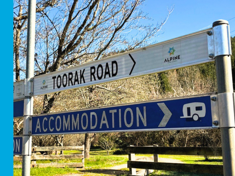 A white street sign with Toorak Road in black lettering and an arrow pointing to the right. A blue sign underneath says 'Accommodation' in white writing. Trees, grass and a blue sky can be seen in the background.