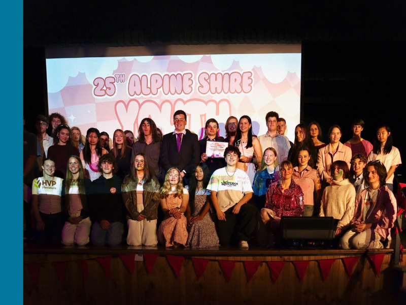 A group of over 20 young people stand or kneel on a stage. There is a project behind them that says 25th Alpine Shire Youth.
