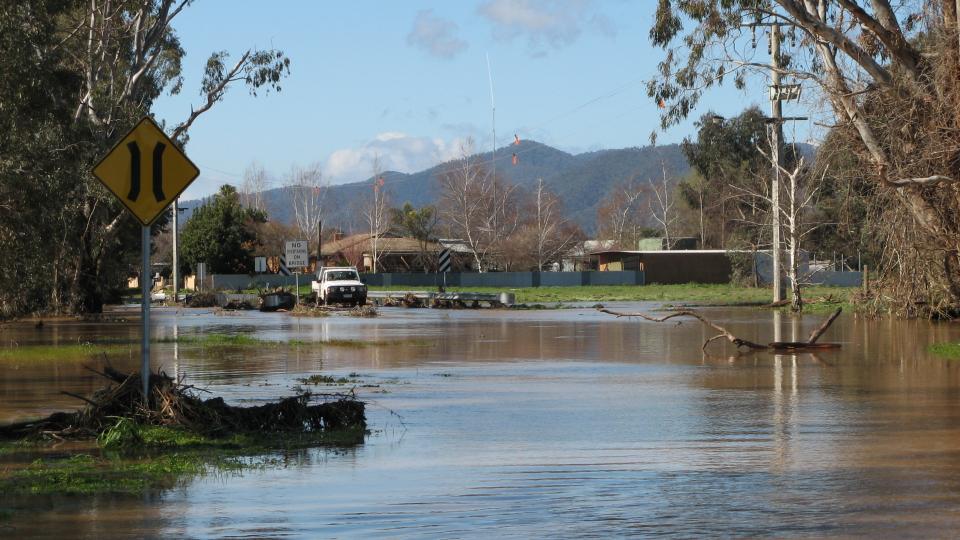 Flood water over a road in Myrtleford. A yellow sign in the forefront indicating it is a bridge, with a white ute on the other side of where the bridge used to be..
