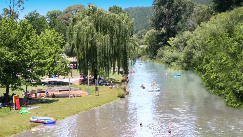 People enjoying themselves in the Ovens River on a sunny day. Inflatables line the green grass on the shores of the river.