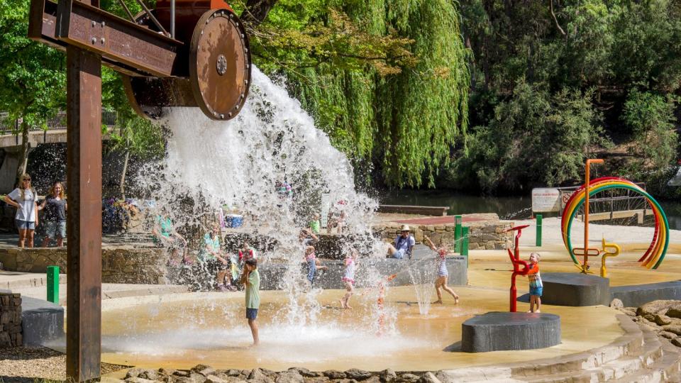 A rust-coloured bucket is dumping water into a splash park, which also has coloured rings for children to run through.