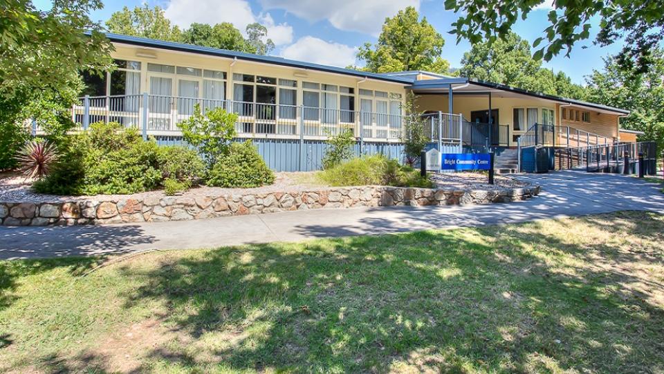 Bright Community Centre is a cream building with blue trim on the railings and roof. It has glass windows and doors lining one wall, overlooking a balcony. The entryway is accessible by both stairs and a ramp. There is a garden in front of the balcony, with a stone retaining wall.