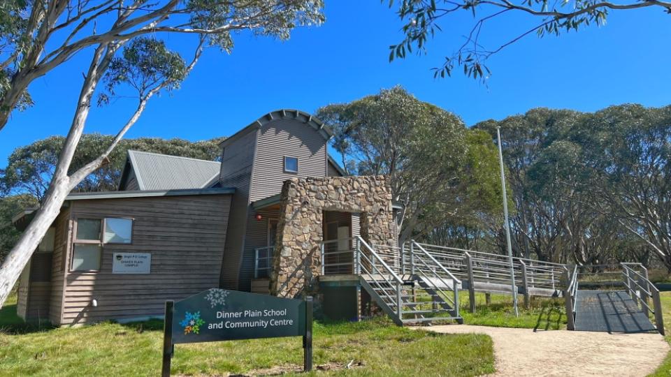 Dinner Plain Community Centre is a two-story building clad in grey weatherboard, corrugated steel, and stone. It has two entrances: stairs and a ramp. There is a blue sign stating Dinner Plain School and Community Centre on the grass to the left of the entrance.