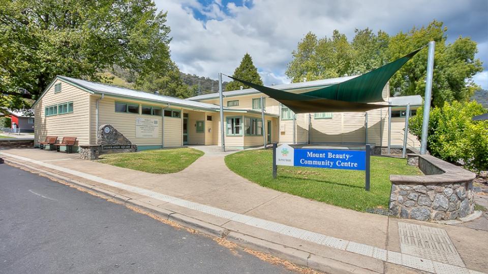 Mount Beauty Community Centre is an L-shaped building clad in cream weatherboard. It has a green lawn in the inner corner of the L with a concrete stage overlooking the lawn. There is a green shadesail covering the lawn. 