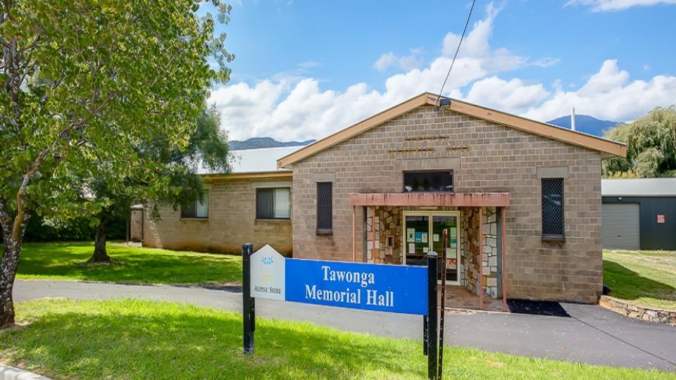 Tawonga Memorial Hall is a brown brick single-story building with a recessed entryway and glass doors. There is a blue sign in front of the building stating Tawonga Memorial Hall. 
