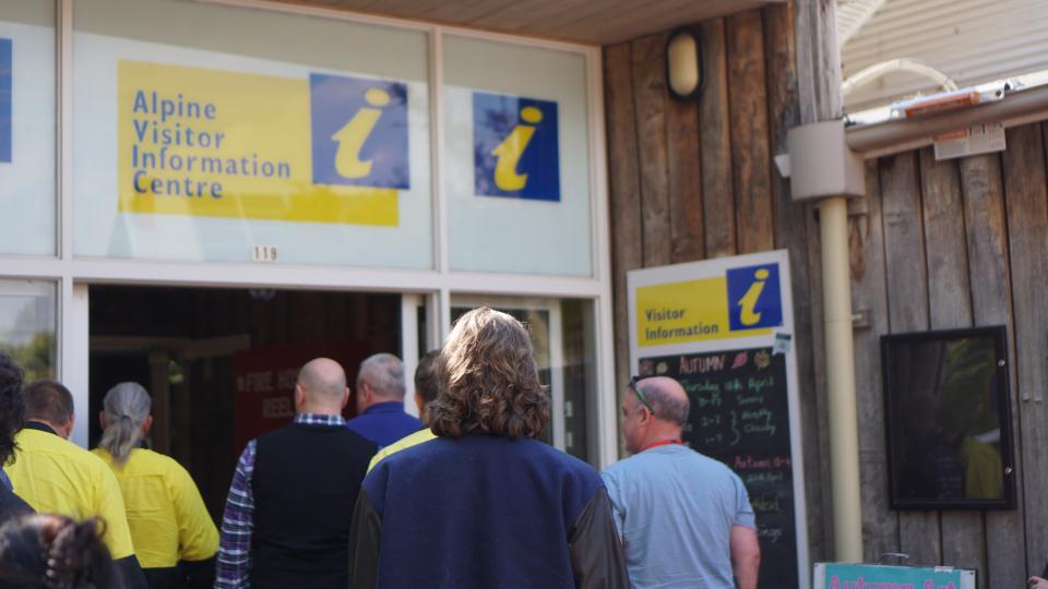 Seven people walk into a building that says Alpine Visitor Information Centre above the doors. We can see the backs of the people walking in.