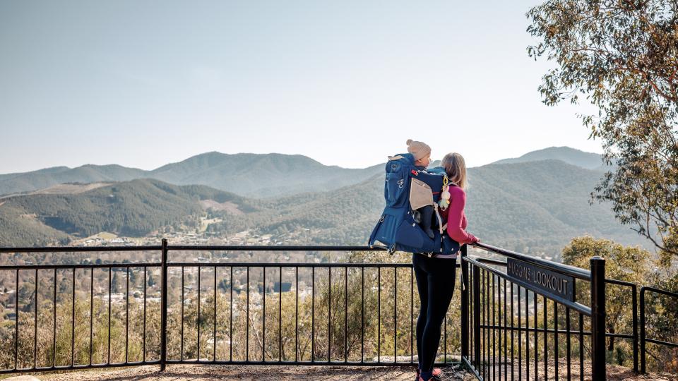 A white woman with blonde hair stands at a lookout, overlooking the town of Bright. She has a baby in a carrier strapped to her back.