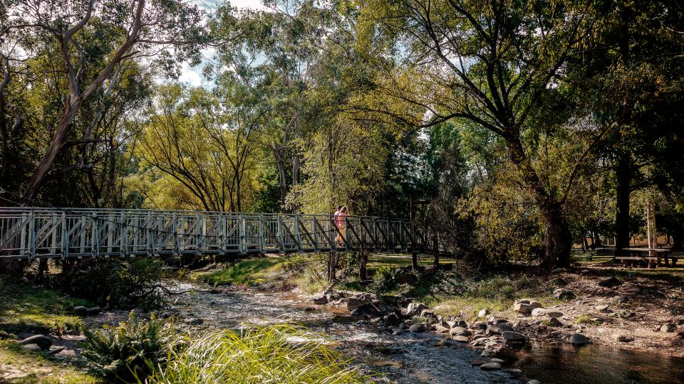 Two people walk across a bridge suspended over a thin river.