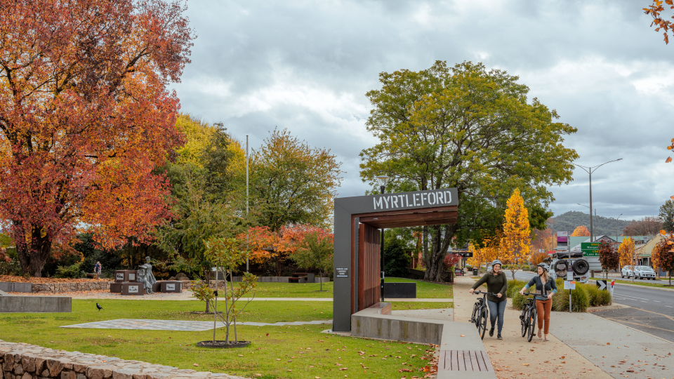 Two women walk their bikes down a footpath alongside the main street of Myrtleford. There is a bus shelter with the word Myrtleford printed along the wooden overhead covering. It is autumn and the trees are vibrant shades of red, orange, yellow, and green.