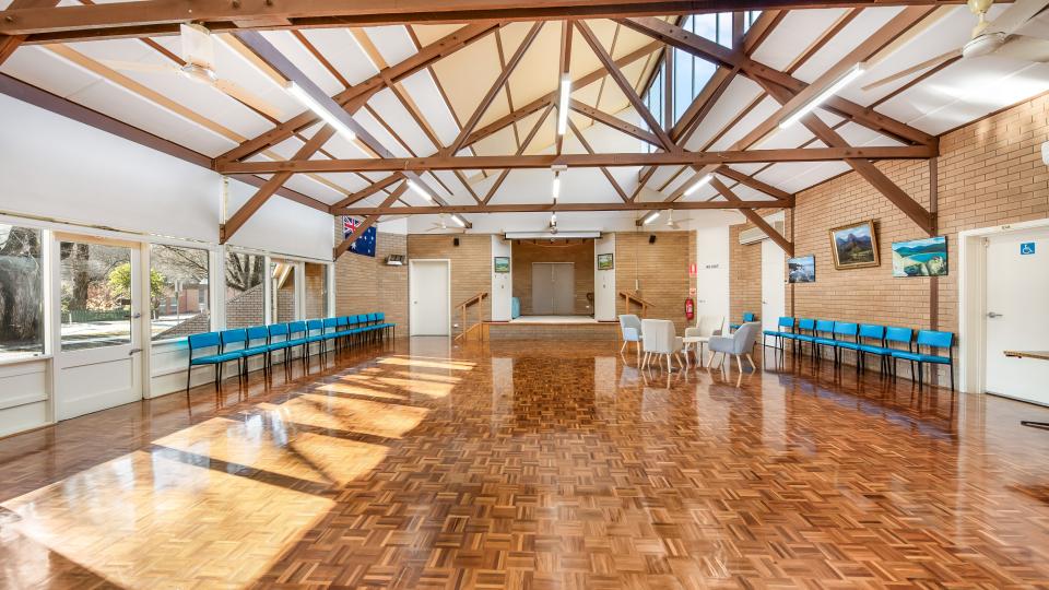 Interior of Bright Senior Citizens Centre with parquet flooring and wooden beams overhead. There are blue chairs lined up on both sides of the room.
