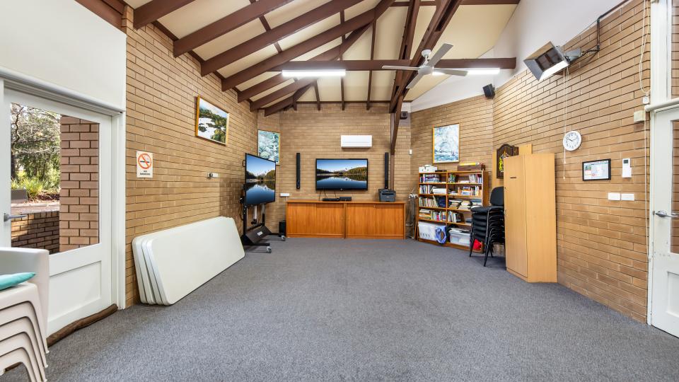 Room inside Bright Senior Citizens Centre with grey carpet and brick walls. There is a wooden buffet cabinet at one end with a television mounted to the wall and another mobile television against another wall. There are folded trestle tables leaning against the same wall as the mobile television and stacks of chairs arranged neatly against other walls.
