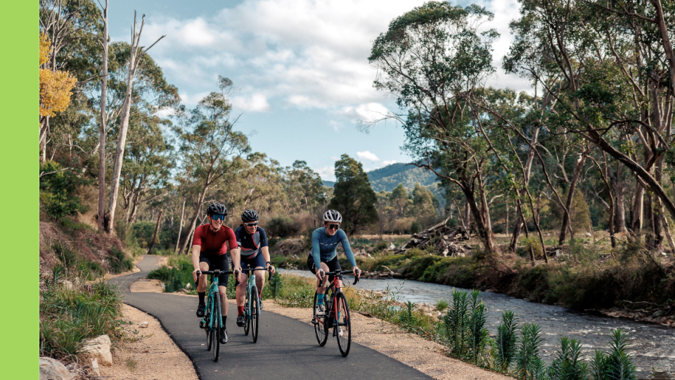 Three cyclists ride along an asphalt riding track next to a river.