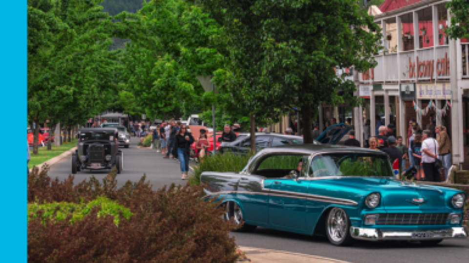 A classic car turquoise and silver in colour drives around a round about in Bright, with a crowd of people walking down the footpath in the background.