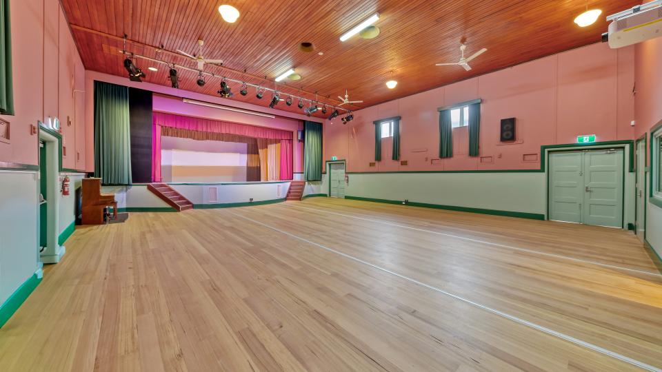 The inside of a community hall with light wooden floors, a stage with pink and green curtains, and green trimming on the walls.