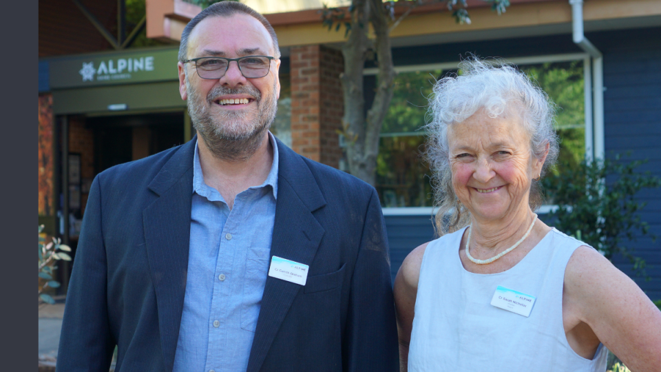 A man wearing a navy blue blazer stands to the left of a woman wearing a pearl necklace and a white linen sleeveless shirt. They are both smiling.
