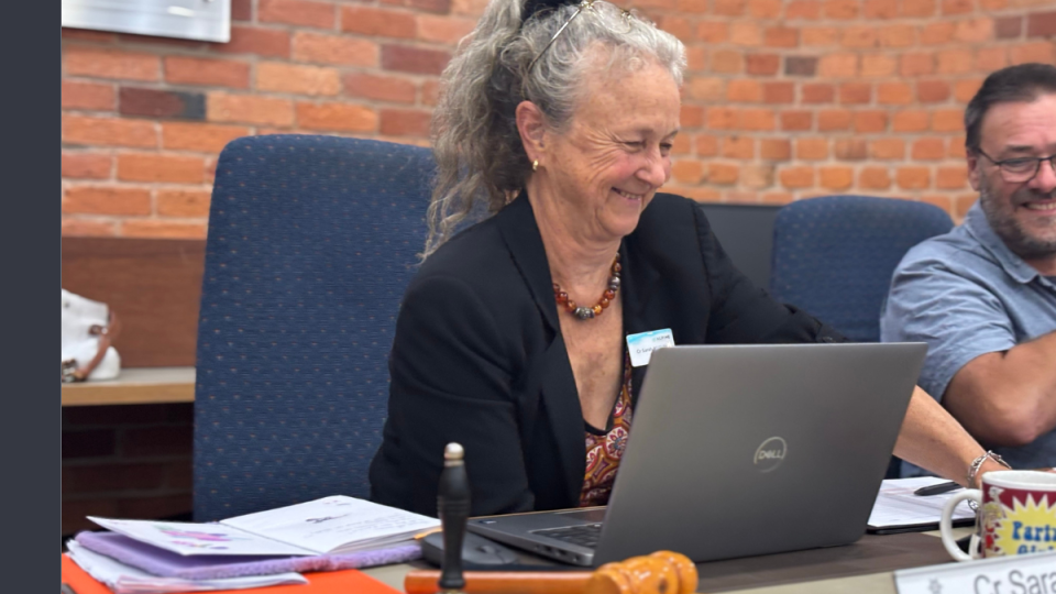 A woman with long grey hair in a high ponytail is smiling while looking down at an open laptop in front of her.