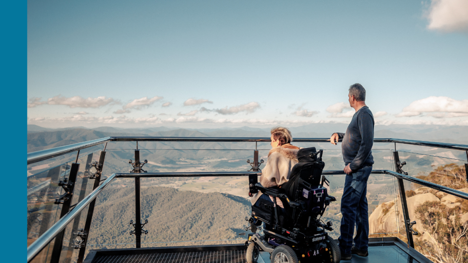 A woman in a motorised wheelchair and a man standing next to her look out over mountains from a lookout platform.