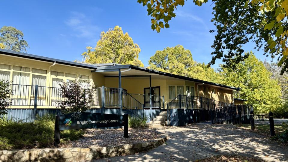 Bright Community Centre is a cream building with blue trim on the railings and roof. It has glass windows and doors lining one wall, overlooking a balcony. The entryway is accessible by both stairs and a ramp. There is a garden in front of the balcony, with a stone retaining wall.