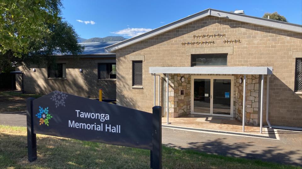 The exterior of the Tawonga Memorial Hall, a cream brick building with a blue sign out the front with its name printed on it.