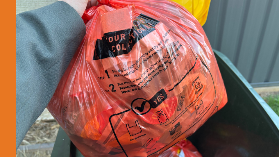 An arm holds an orange plastic bag over an open yellow-lidded kerbside recycling bin.