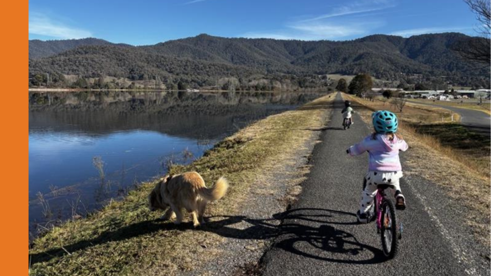 A young child rides a bike on a path next to a lake. There is a Golden Retriever dog next to her.