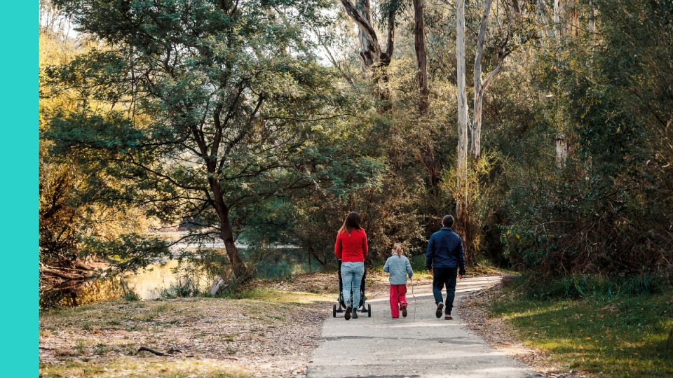 Three people, including a woman, a child, and a man, walking along a footpath. The woman is pushing a pram. We can only see their backs. There is lots of greenery in the background, suggesting a bush setting.