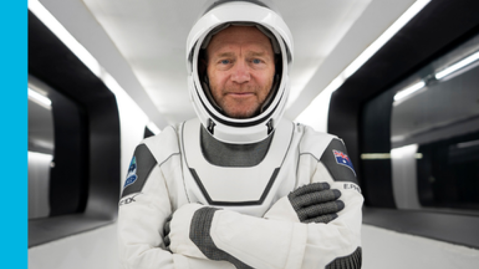 Eric Philips stands in a space craft with his crossed looking directly at the camera wearing a space suit, including helmet.