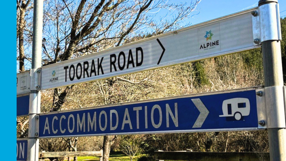 A white street sign with Toorak Road in black lettering and an arrow pointing to the right. A blue sign underneath says 'Accommodation' in white writing. Trees, grass and a blue sky can be seen in the background.